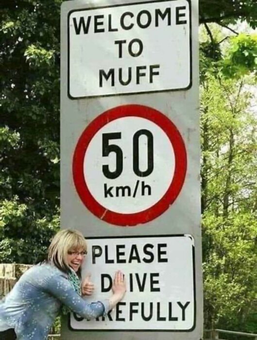 Kvinna poserar leende vid vägskylt "WELCOME TO MUFF", hastighetsgräns 50 km/h och "PLEASE DRIVE CAREFULLY".