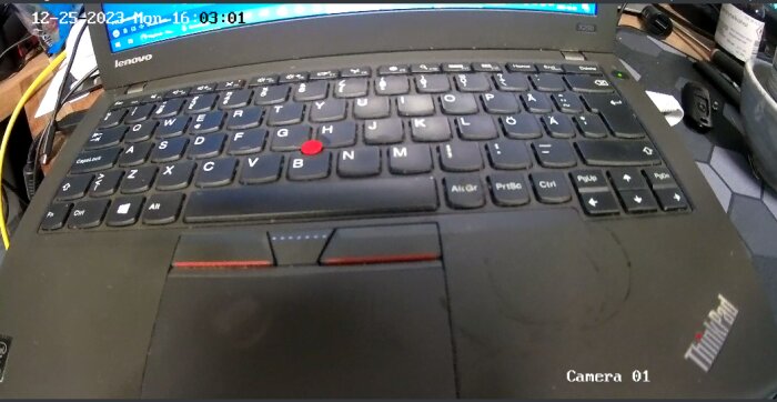 Svart Lenovo ThinkPad laptop, tangentbord, TrackPoint, slitna handledsstöd, skrivbordsbakgrund, säkerhetskamera vy, datumstämpel.