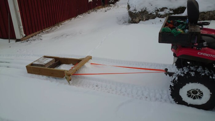 Fyrhjuling kopplad till träsläde på snö, röd lada, vinter, utomhus, draglina, arbetsfordon, kallt väder.