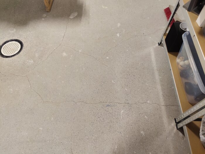 Ett garagegolv med sprickor, kvast, förvaringslådor och en golvbrunn.