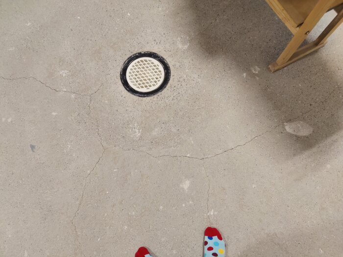 Golv med golvbrunn, sprickor, trästolens ben, person i färgglada sockor.
