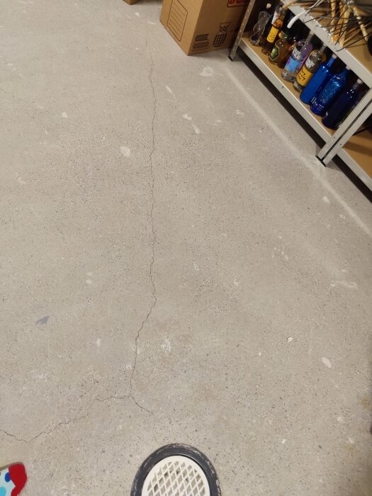 Sprucken golvplatta inomhus, hyllor med flaskor, kartonglåda, rund golvbrunn.