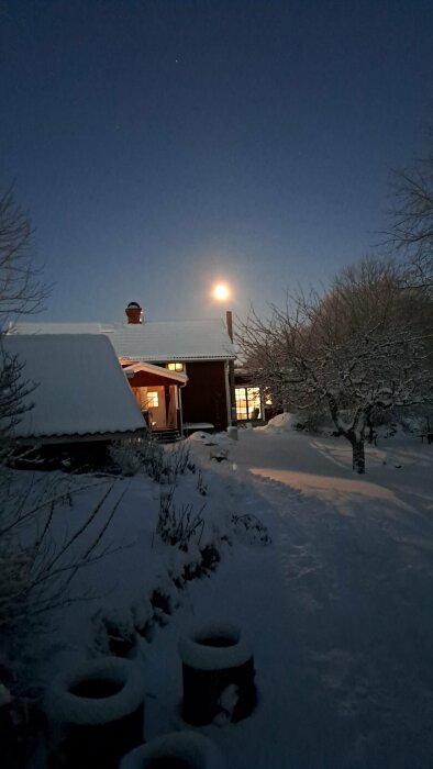 Ett snöigt landskap vid skymning med hus, träd, och fullmåne som lyser upp himlen.