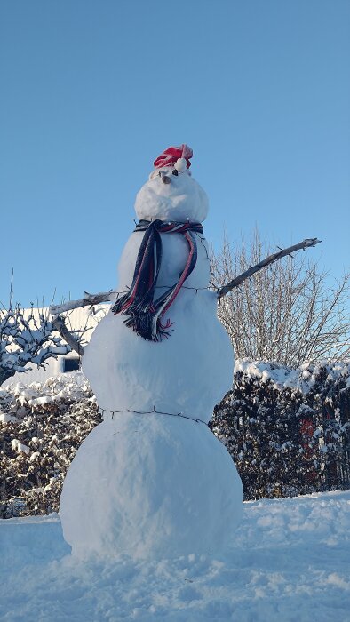Stor snögubbe med hatt, scarf, grenarmar och snöig bakgrund under klarblå himmel.