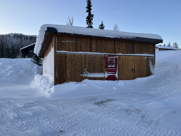 Trästuga med röd dörr, omgiven av snö, vinterlandskap, klar himmel, snötäckt tak, dagtid.