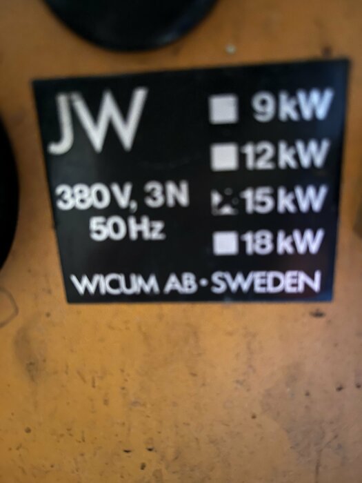 Svart skylt med vit text, strömspecifikationer, suddig bakgrund. Märkt "JW", "WICUM AB - SWEDEN", olika kilowattvärden.