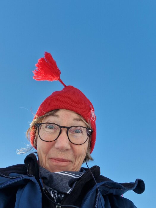 Person med röd mössa och tofs, glasögon, utomhus, blå himmel, tar selfie, ser lycklig och överraskad ut.
