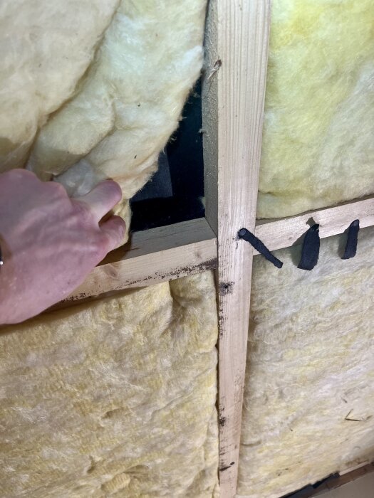 En hand pekar på en tom plats i en väggisolering av glasull mellan träreglar.