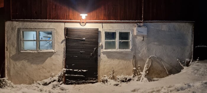 Vitputsat hus med snö, träport, fönster, upplyst av taklampa på kvällen.