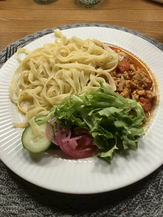 Tallrik med pasta, köttfärssås, grönsallad, gurka, picklad rödlök på träbord.