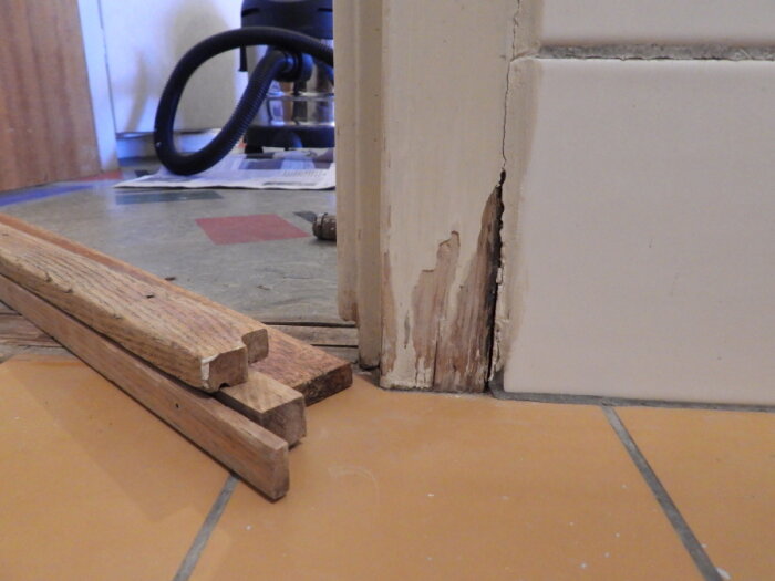 Gulvfliser, skadet dørkarm, trälist, dammsugarslang bakom, tecken på renovering eller reparation.