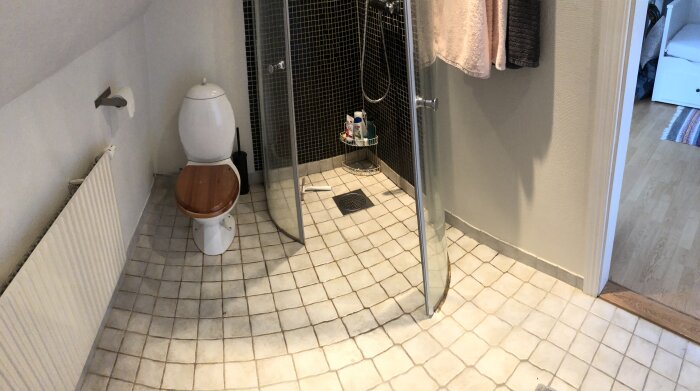 Ett badrum med toalett och duschhörna, kakelgolv, trätoalettsits och handdukar.