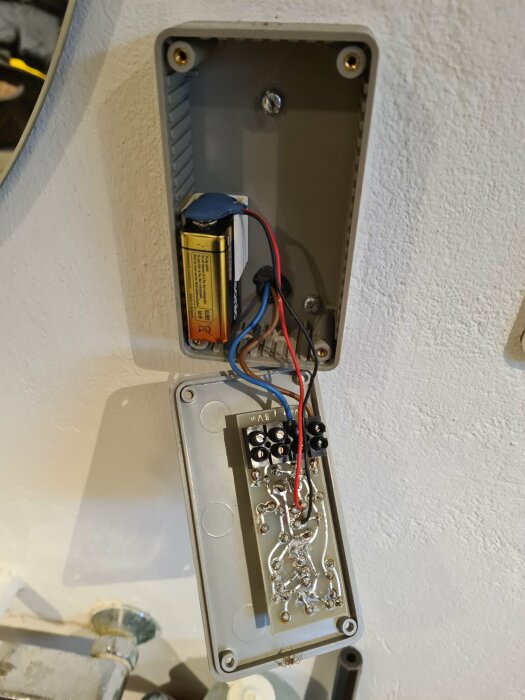 Öppen elektroniklåda på vägg med batteri och kretskort, oavslutad installation eller reparation.
