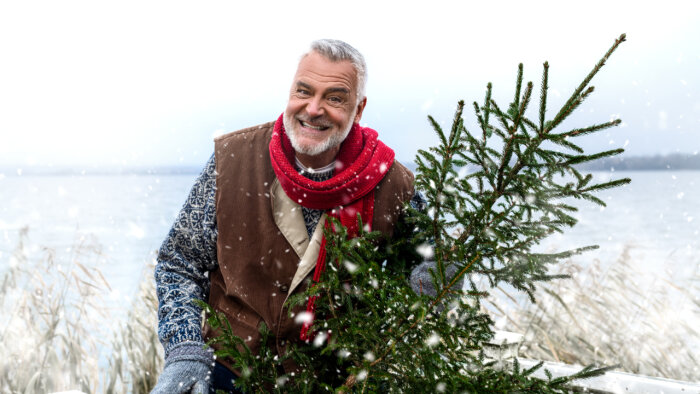 Leende man håller julgran utomhus i snöigt vinterlandskap.