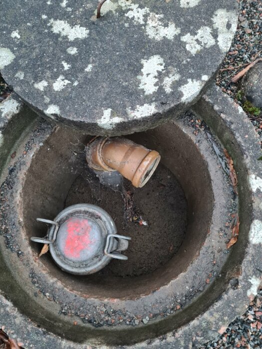 Ett öppnat brunnslock visar en brunnskammare med ett rör och spindelväv, omgivet av mossa och småstenar.