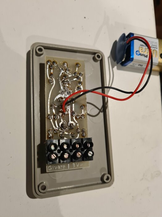 Elektronisk krets med lödningar, terminalblock och batteri på vit yta, etikett märkt "Givare 3V".