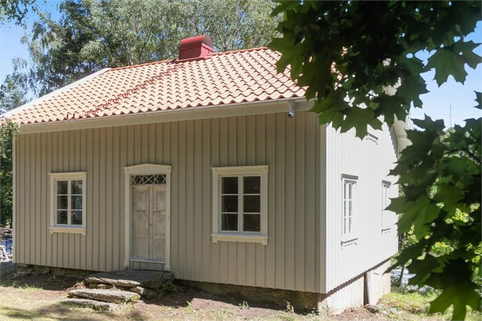 Ett litet traditionellt hus med röda takpannor, vitmålad träfasad, grönskande träd och stensteg framför dörren.