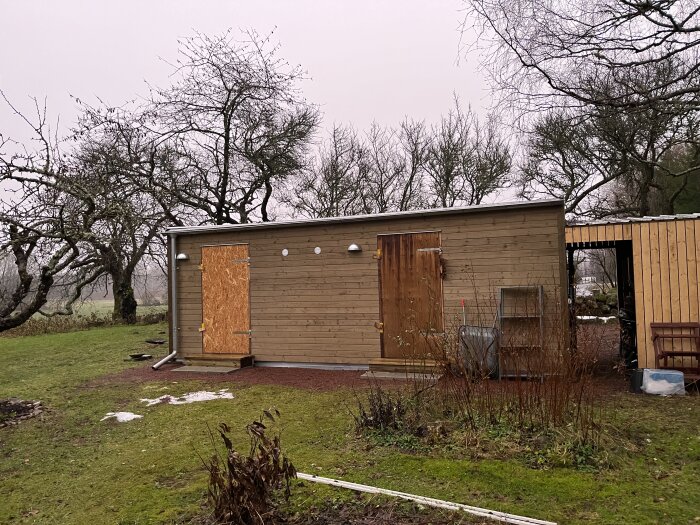 Trädgårdsskjul, trästaket, kala träd, grå himmel, rester av snö, kallt väder, övergivna möbler, landsbygdsmiljö.