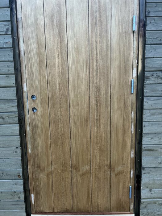 En träfärgad dörr med gångjärn, lås och handtag, placerad mot en träbeklädd vägg.