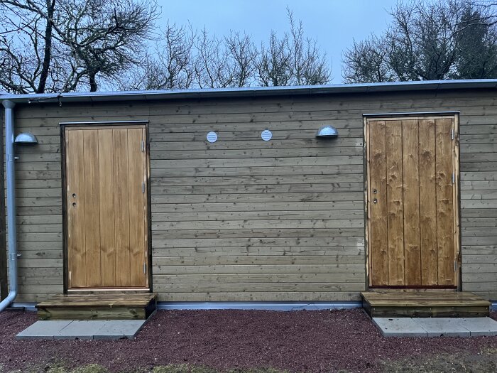 Träbyggnad med två dörrar, belysning ovanför, rödaktigt underlag, träd i bakgrunden.