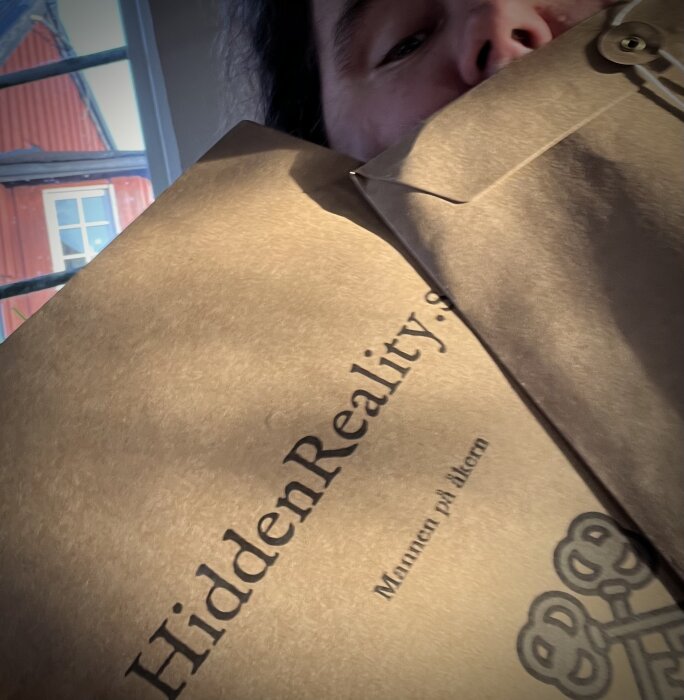 Person håller en öppen bok, titel "Hidden Reality", närbild, selektivt fokus, delar av ansikte synliga.