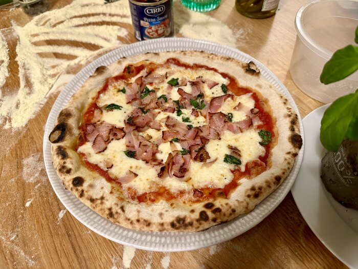 Nybakad pizza med tomatsås, ost och skinka på ett mjölad bord med tomatsåsburk och basilika.