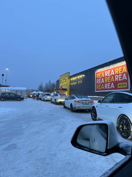 Parkering vid köpcentrum på vintern, skylt med "REA", bilar i kö, snöig mark.