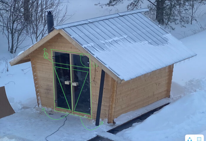 En trästuga med snötak och istappar, omgiven av snö, med markerade konturer och pilar på bild.