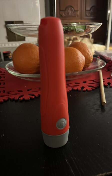 Röd stavmixer framför apelsiner på glasfat med röd duk på svart bord.