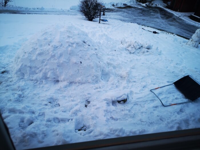 Stor snöhög framför fönster, spade, snöig mark, vinterväder, kallt, dagtid, väg i bakgrunden.