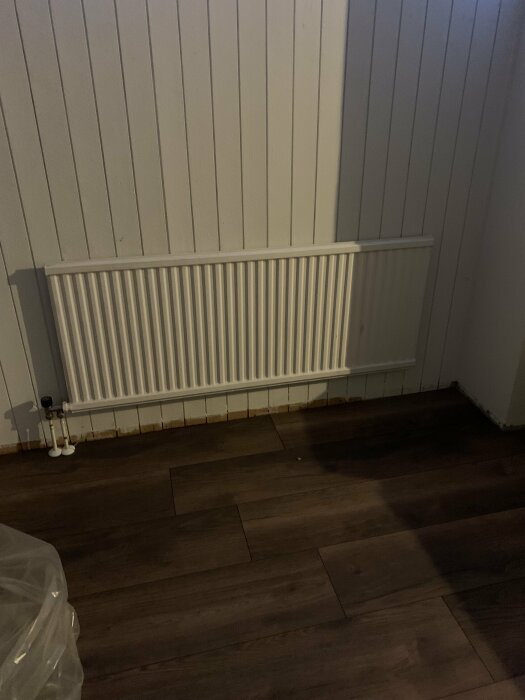 Väggmonterad radiator i rum med panelvägg och mörkt laminatgolv. Opersonlig, enkel, inredningsdetalj.