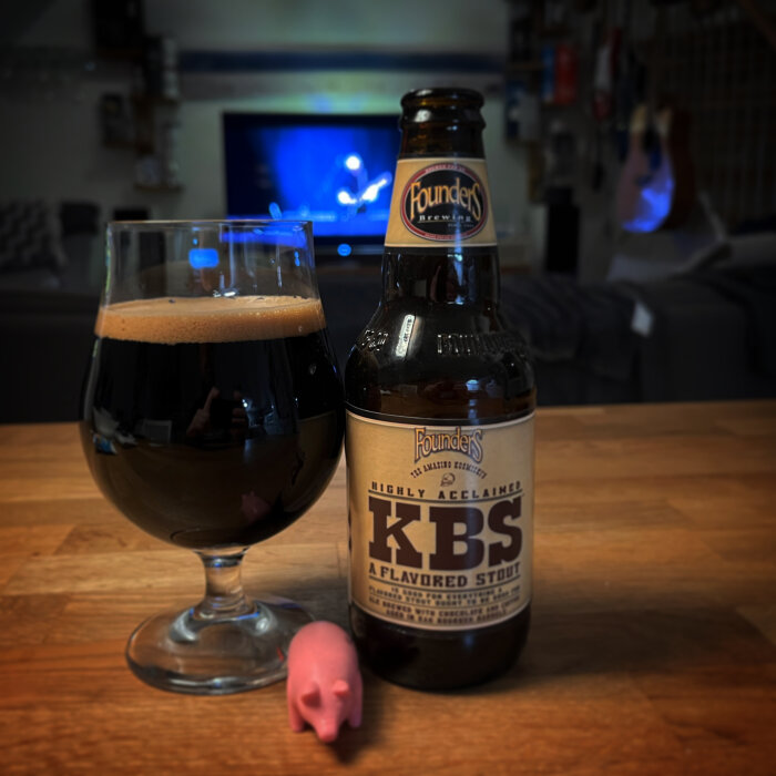 Stout i glas, ölflaska, liten rosa figur, suddig TV i bakgrunden, inne, mysig atmosfär.