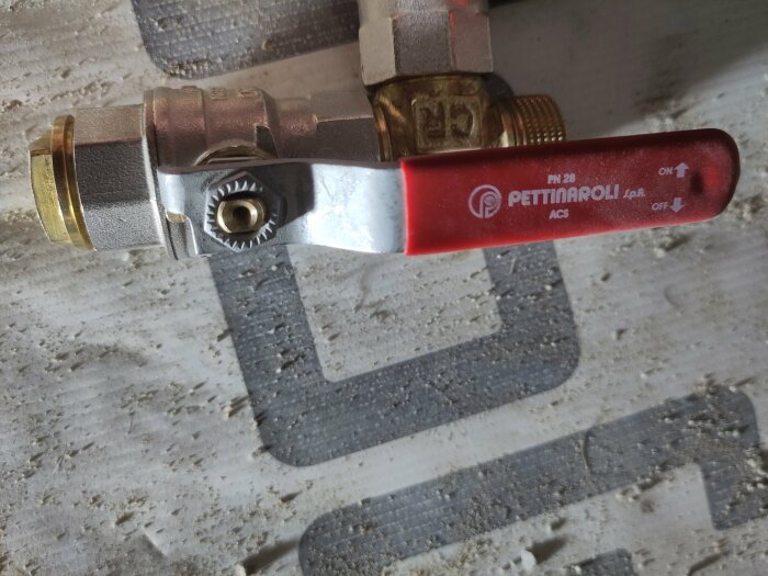 Röd kulkran för VVS-installation mot betongvägg, dammig miljö, Pettinaroli-märke, rörkopplingar i förgrunden.