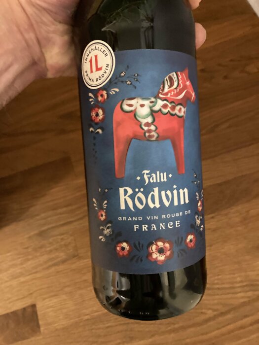 Flaska med etikett 'Falu Rödvin', fransk rött vin, dalahästmotiv och blommor, hålls i hand.