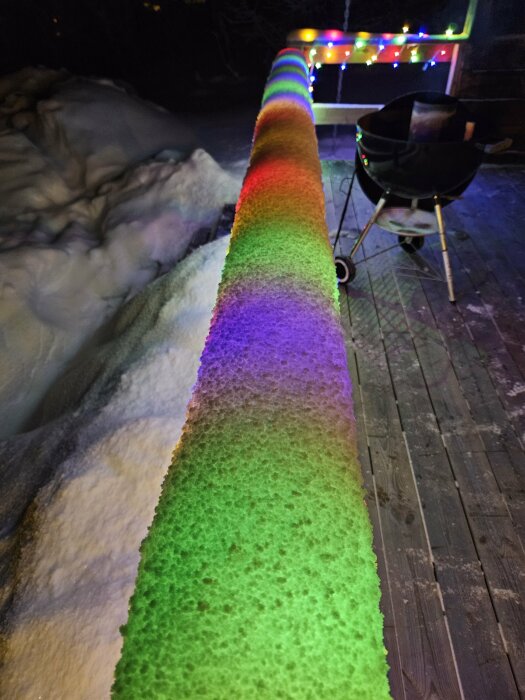 Snö täckt räcke med regnbågsreflektion; brokiga lampor i bakgrunden; grill; kväll; vinter.