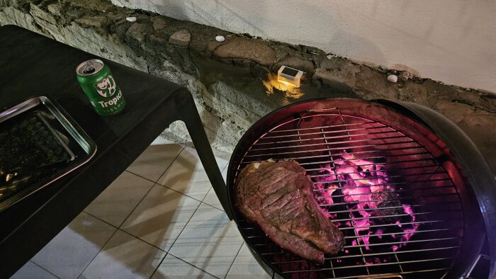 Kvällsgrillning utomhus, kött på grillen, öppen burk öl på bordet, mysig belysning, avkopplande stämning.