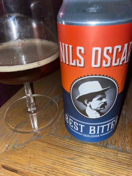 Ölburk märkt "NILS OSCAR", "BEST BITTER" bredvid glas med mörkt öl, träyta.