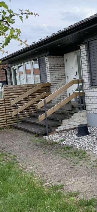 Ett svart hus med terrass, trätrappa, vit dörr och tegeldetaljer. Nedfallna löv och grön gräsmatta syns.