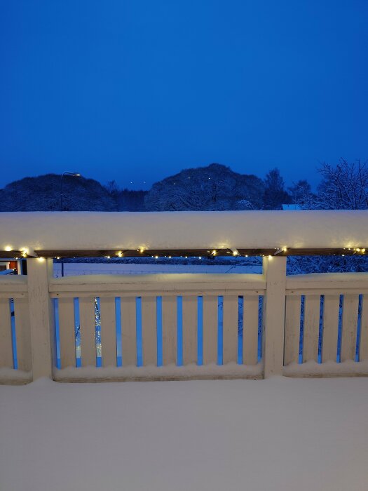 Snötäckt altanräcke, ljusslinga, skymning, frostiga träd, lugnt vinterlandskap.