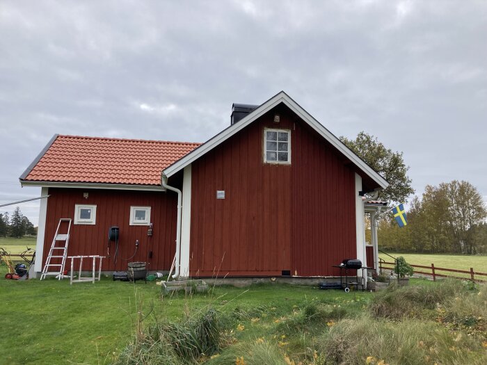 Traditionellt rött svenskt hus med vita knutar, trädgård, svensk flagga och stege vid sidan.