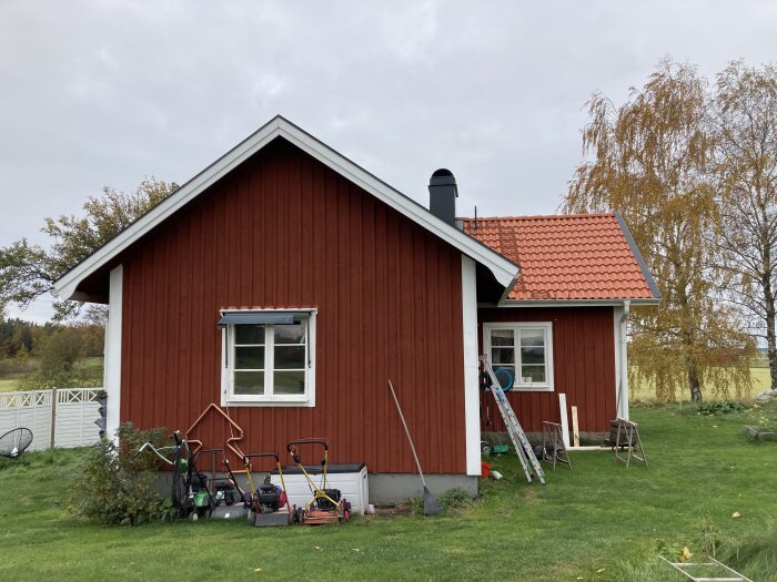 Rött hus med vita knutar, gräsklippare framför, grönska, höst, stege, molnig himmel, lugnt bostadsområde.