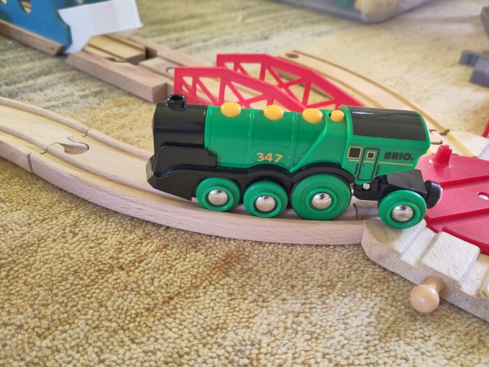 Grön, svart BRIO leksakståg på träjärnvägsspår, röd bro, matta, barnleksaker.