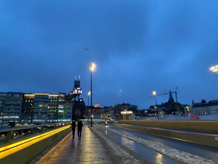 Kvällsutsikt över en bro, människor som promenerar, upplysta stadsskyltar och byggnader, mörkblå himmel.