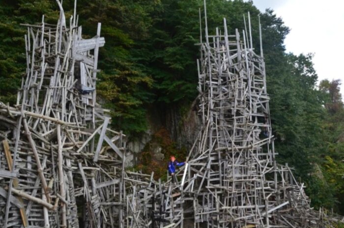 Komplex träkonstruktion i skog, person klättrar, konstnärlig, stor, improviserad arkitektur mot gröna träd.