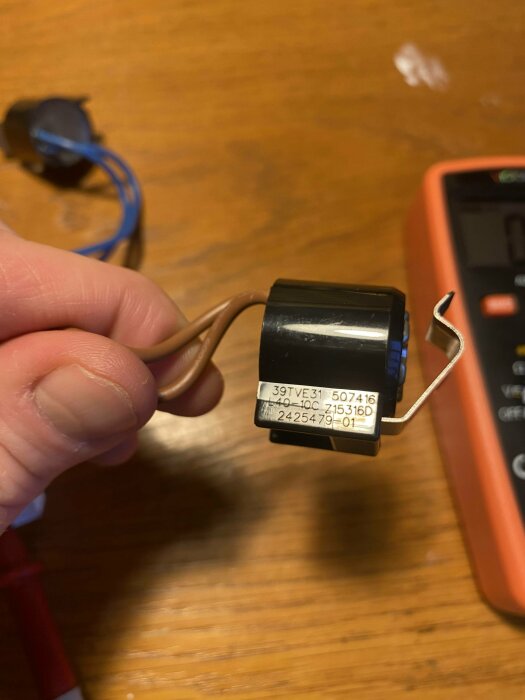 En hand som håller en elektronisk komponent med kablar nära ett multimeter på träbord.