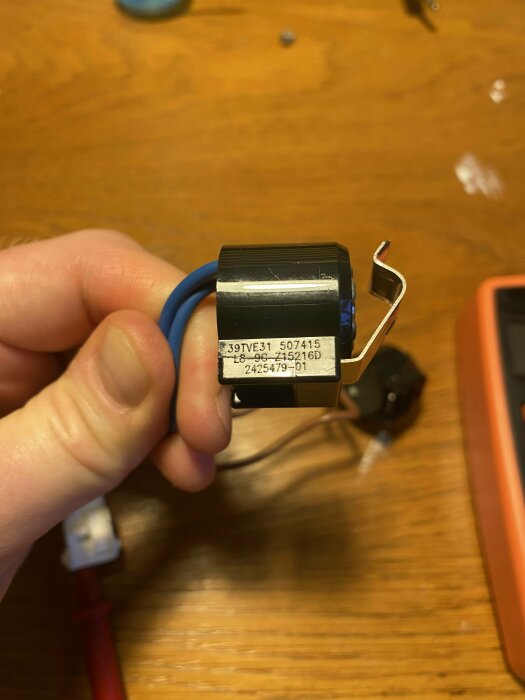 Hand håller en elektrisk komponent med kablar mot ett träbord. Etiketter och siffror synliga på komponenten.