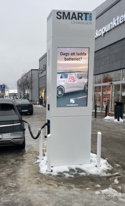 En laddstation för elbilar märkt med "SMART Charger", vinterväder, QR-kod, bil i bakgrunden.