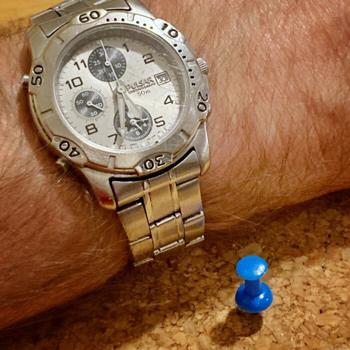 Silverfärgad armbandsklocka på handled, blå häftstift nära. Urtavla med kronograf. Tidmätning, mode, tillbehör.