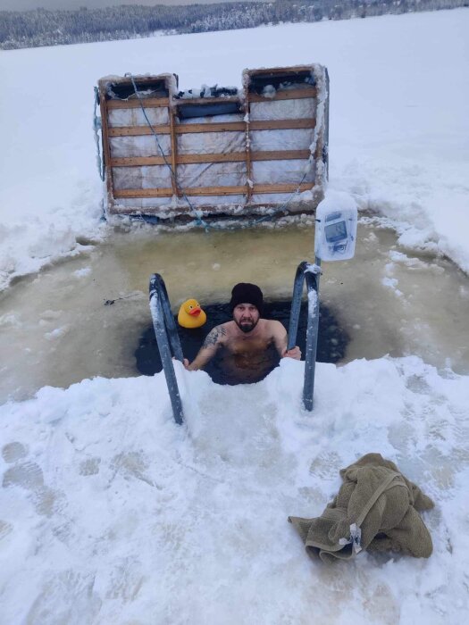 Person isbadar i hål på frusen sjö, med badanka, stege, handduk och isiga träd i bakgrunden.