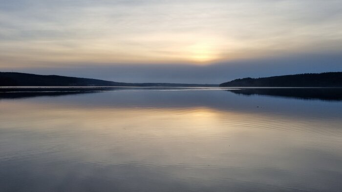 Solnedgång över stilla vatten, reflekterande ljus, skogbeklädda kullar i bakgrunden, fridfullt landskap.